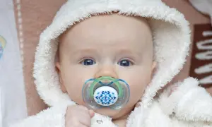 Söt snorig bebis med blå ögon och en napp med mönster, klädd i en vit fluffig mössa.