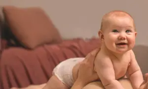 Glad bebis i blöja, kanske med tandsprickning, tittar uppåt. En hand håller om barnet.
