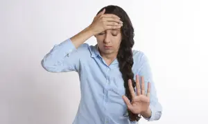 Kvinna med handen mot pannan, ser ut att ha ont i huvudet, kanske gravid. Hon håller upp en hand som för att stoppa något.