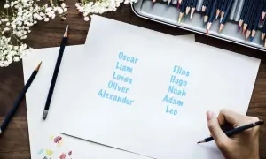 En hand skriver fina pojknamn som Oscar, Liam, Elias och Hugo på papper. Färgpennor och blommor ligger på träbordet.