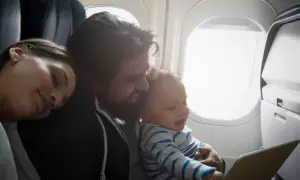Familj flyger med bebis. Pappan håller barnet som tittar på en surfplatta medan mamman sover.