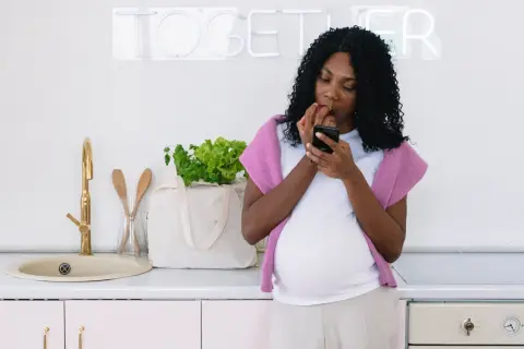 Gravid kvinna i köket kollar mobilen och äter ett äpple, en påminnelse om vad man ska undvika som gravid.