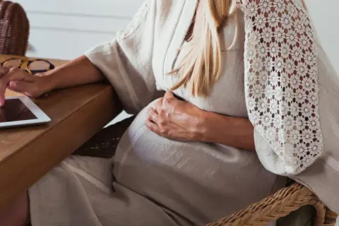 Gravid kvinna med handen på magen, använder en surfplatta vid ett träbord.