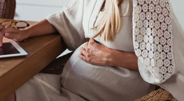 Gravid kvinna med handen på magen, använder en surfplatta vid ett träbord.
