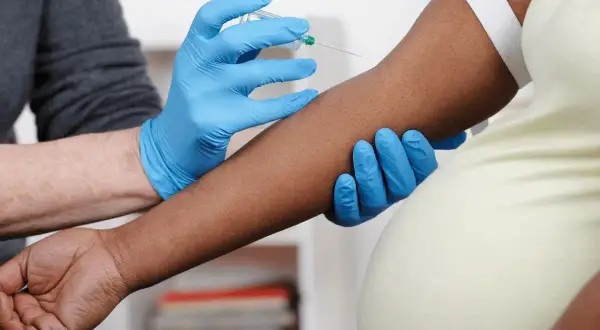 Blodprov tas för nipt test vecka 10. En hand med blå handske håller armen medan en annan hand med blå handske för in en nål.
