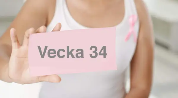 En person håller upp en rosa lapp med texten "Vecka 34". En rosa band pryder personens vita linne.