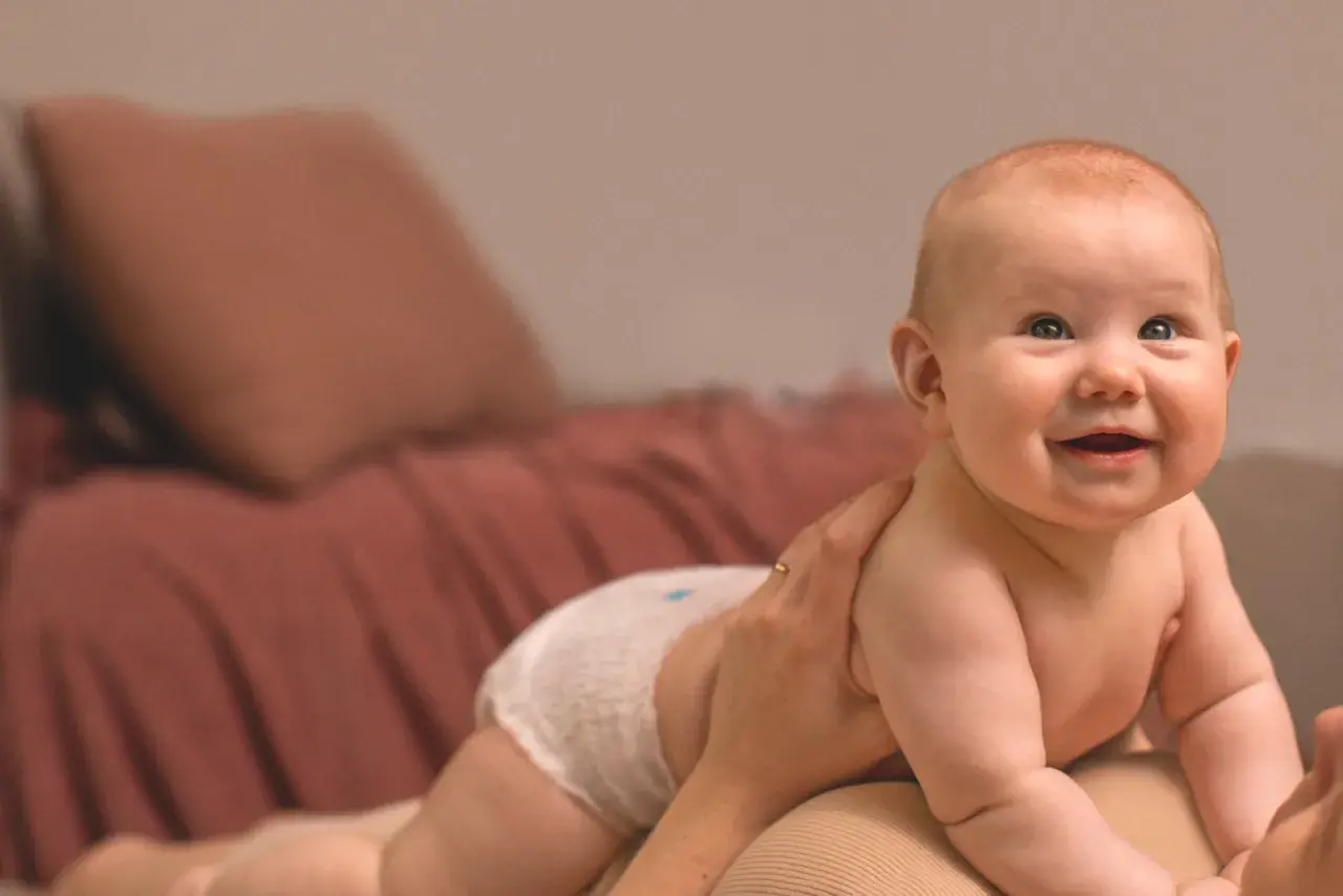 Glad bebis i blöja, kanske med tandsprickning, tittar uppåt. En hand håller om barnet.