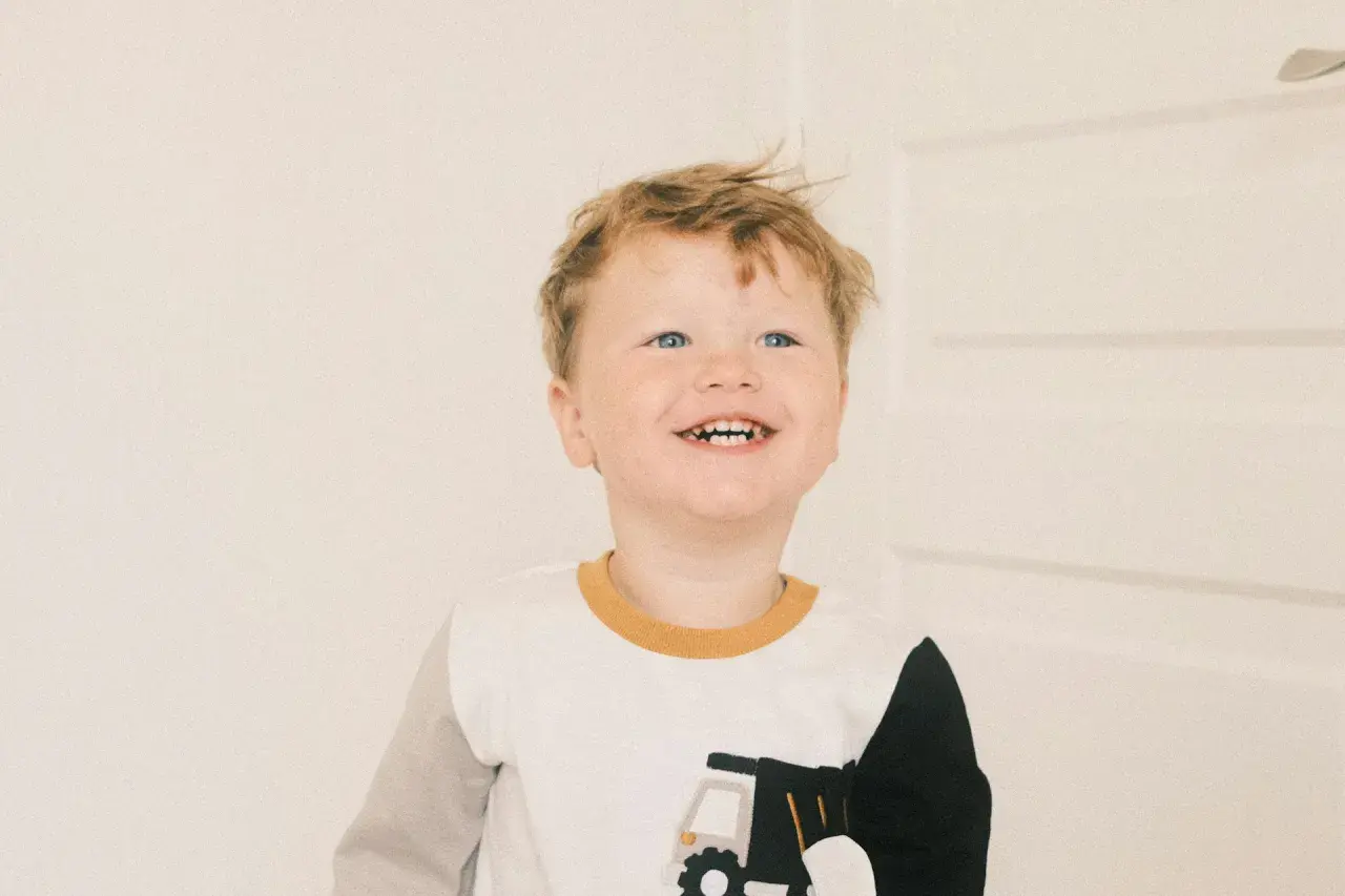 Glad pojke med blont hår och blå ögon ler mot kameran. Han har på sig en tröja med en lastbil på. Han är det andra barnet i familjen.