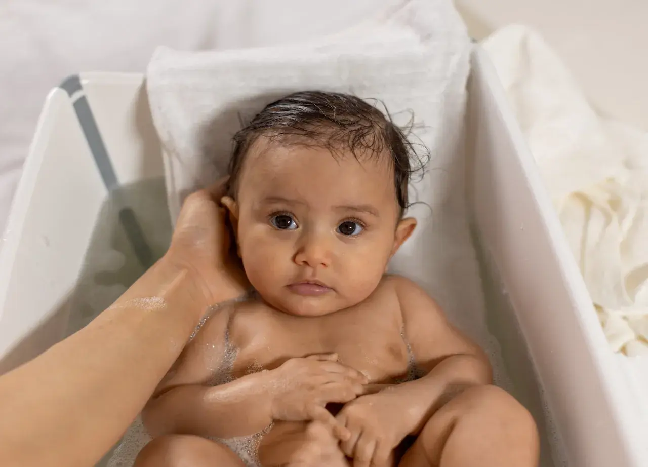 Söt bebis i badet, med lite skorv i håret. En hand tvättar försiktigt.