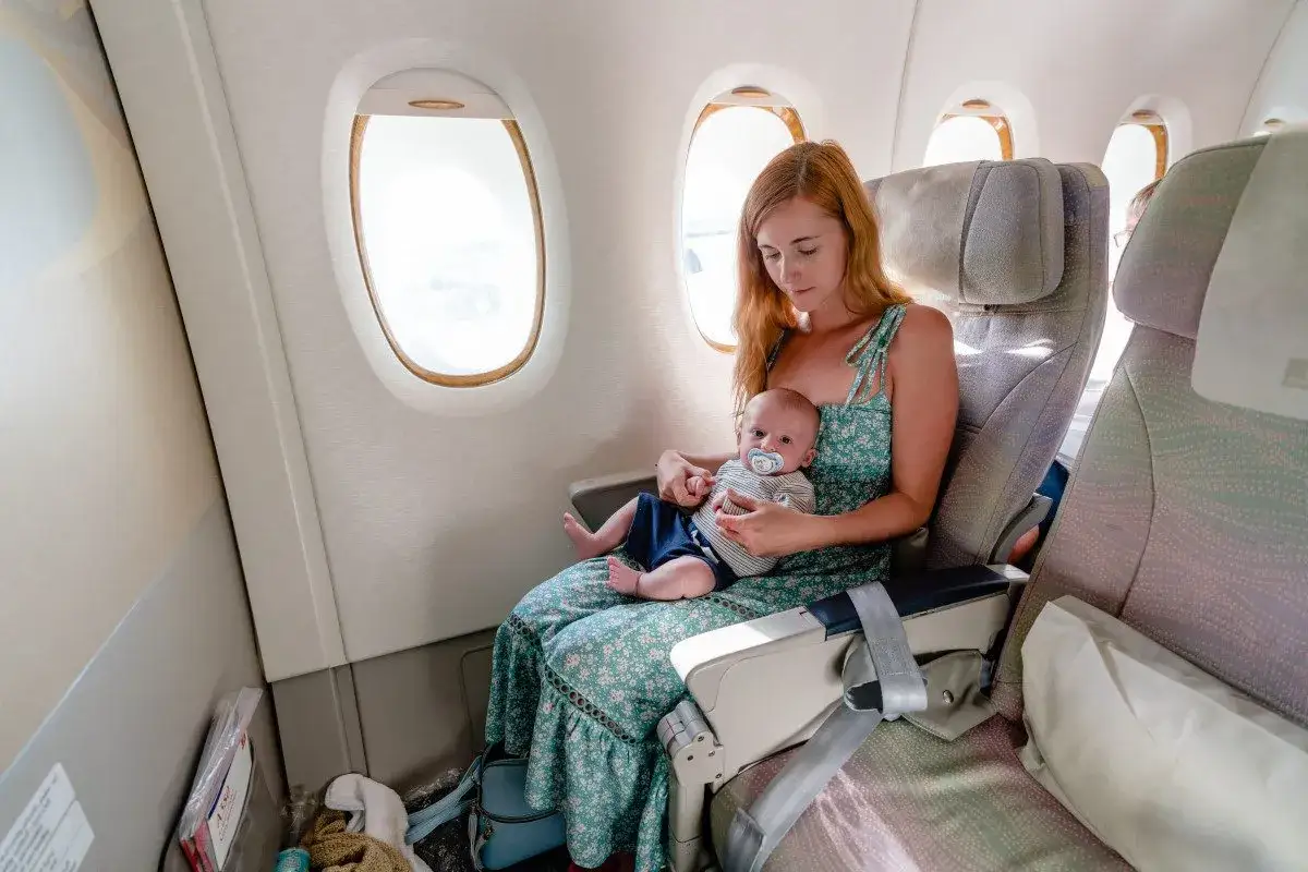 En kvinna med rött hår sitter vid ett flygplansfönster och håller en bebis i famnen. De är på väg att flyga med bebis.