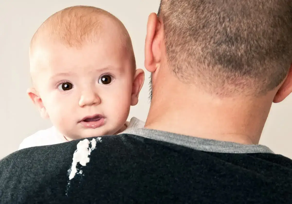 En s&ouml;t bebis med stora &ouml;gon tittar fram &ouml;ver pappans axel, som har f&aring;tt en liten kladd av mat p&aring; sig.