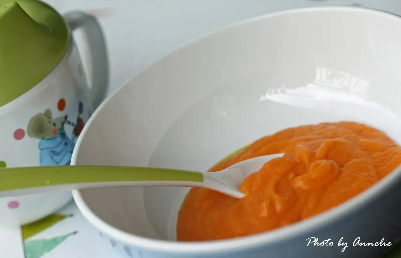En skål med morotspure bebismat, redo att matas. En grön sked ligger i purén.
