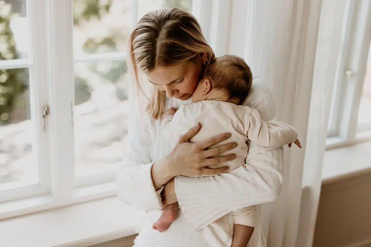 A mother holds her baby close, both wearing light-colored clothing, by a window.