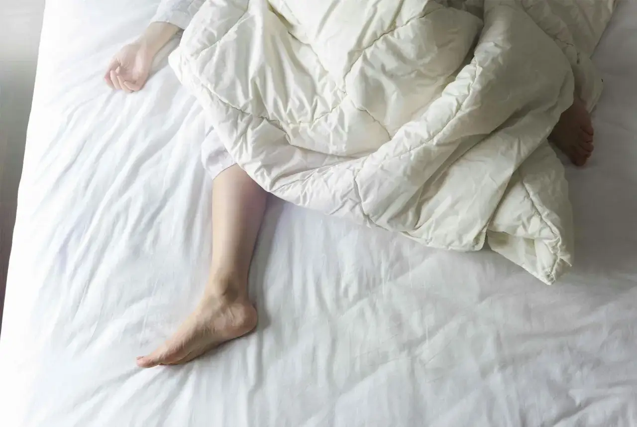 En person sover i en vit säng, täckt av ett vitt täcke. En arm och ett ben syns. Kanske upplever personen svettningar gravid.