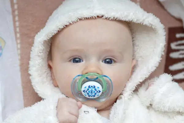 Söt snorig bebis med blå ögon och en napp med mönster, klädd i en vit fluffig mössa.