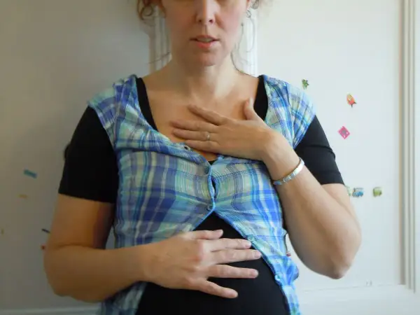 En kvinna med en rutig skjorta över en svart topp, håller händerna över magen. Hon ser ut att vara **rennie gravid**.