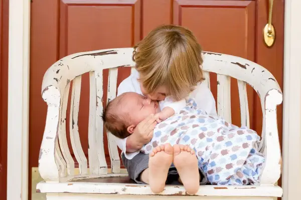 Äldre syskonet kysser sin nyfödda bebis på en sliten bänk. En vacker stund att få syskon.