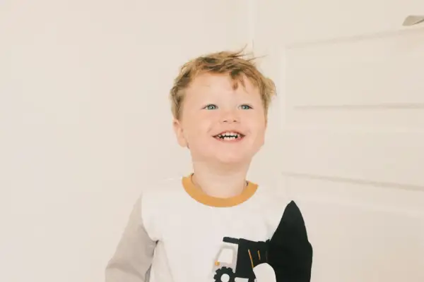 Glad pojke med blont hår och blå ögon ler mot kameran. Han har på sig en tröja med en lastbil på. Han är det andra barnet i familjen.