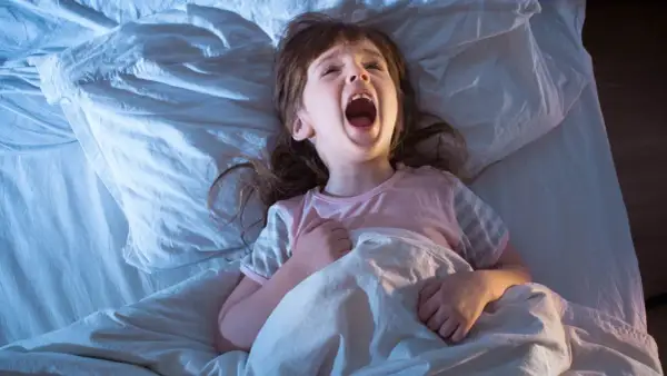 Ett litet barn vaknar och skriker hysteriskt i sängen, med öppen mun och uppspärrade ögon.