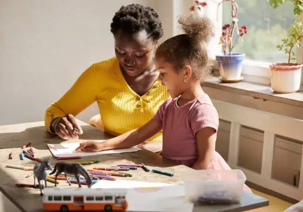 Ensamstående mamma och dotter pysslar vid ett bord med kritor och leksaker.
