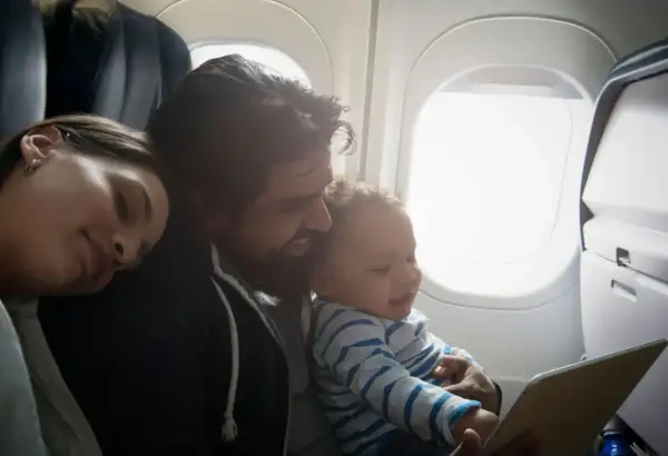 Familj flyger med bebis. Pappan håller barnet som tittar på en surfplatta medan mamman sover.